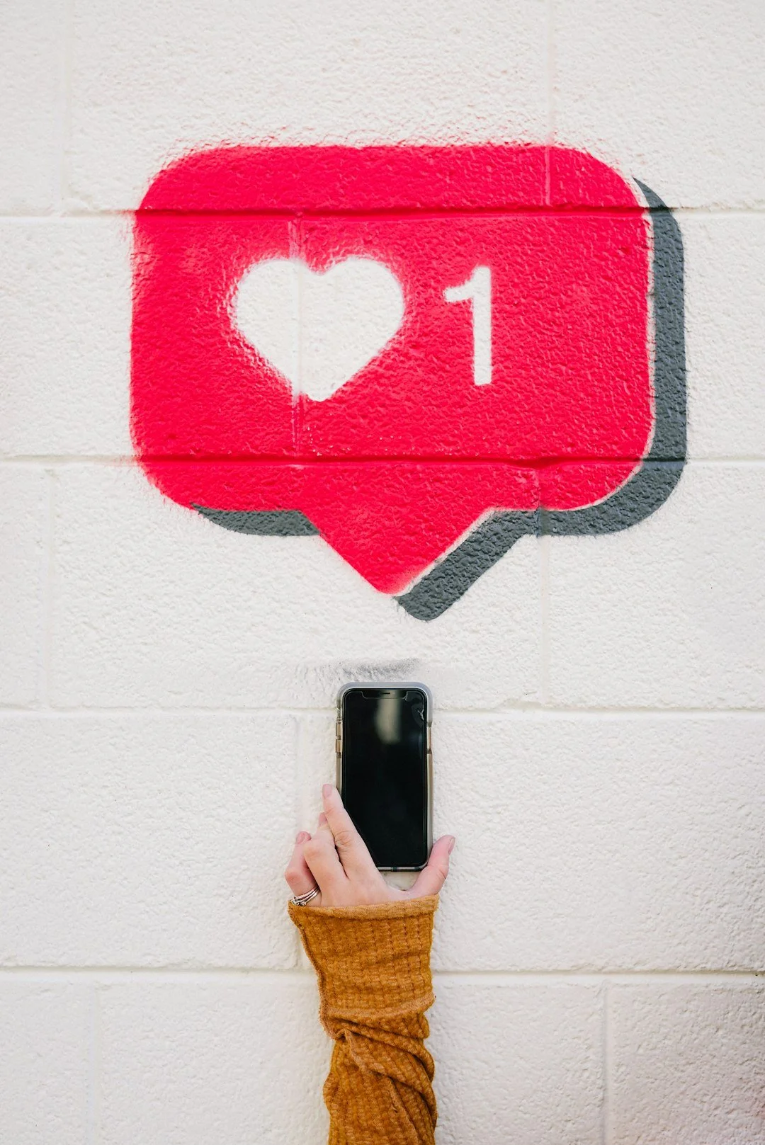 A hand holding a phone below a large painted social-media like icon on a white wall